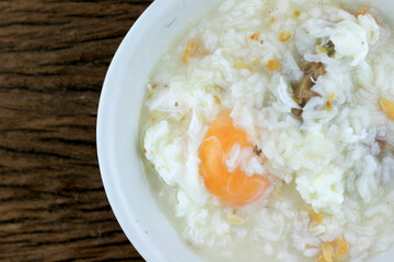 Homemade Thai Rice Soup With chicken, pork and Soft boiled egg or onsen egg in white blow on rustic wooden background. Thai style Food. Top view.