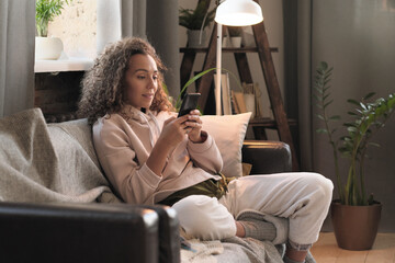 Fototapeta premium Young blogger sitting on sofa and working online on her mobile phone at home