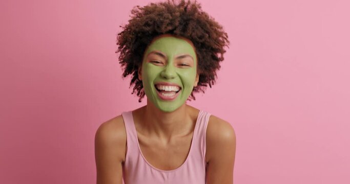 Serious young African American woman appears suddently from bottom of pink background raises palms and says boo then smiles joyfully has curly bushy hair applies green beauty mask for skin care