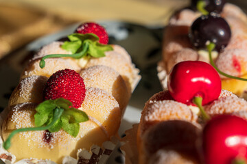 Artificial desserts presenting realistic sponge cakes coated with sugar with cherries and berries on top wrapped in baking paper, on a white glass tray with black ornaments during golden hour
