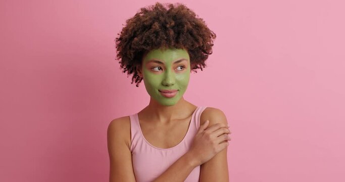 Gorgoeus African American woman looks tenderly at camera has tender calm expression applies green mask on face for flaw perect skin dressed casually poses against rosy background. Face care concept