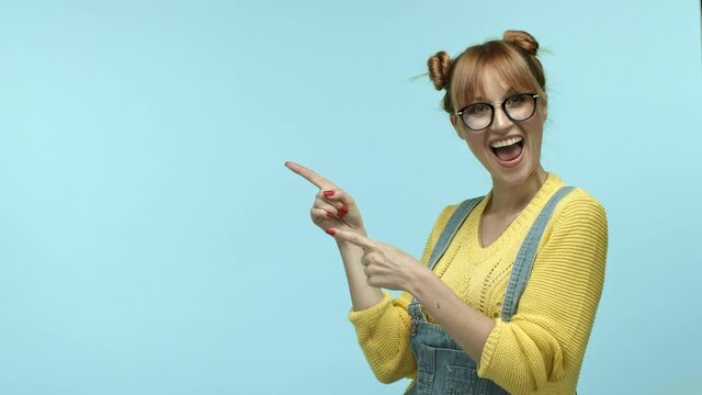 Cheerful girl hipster in glasses, with cute bangs and buns hairstyle, pointing fingers left at copy space for logo or promo offer, showing thumbs-up, recommending special deal, blue background