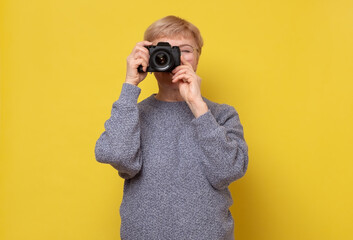 Mature female photographer with photocamera by face standing on yellow background.