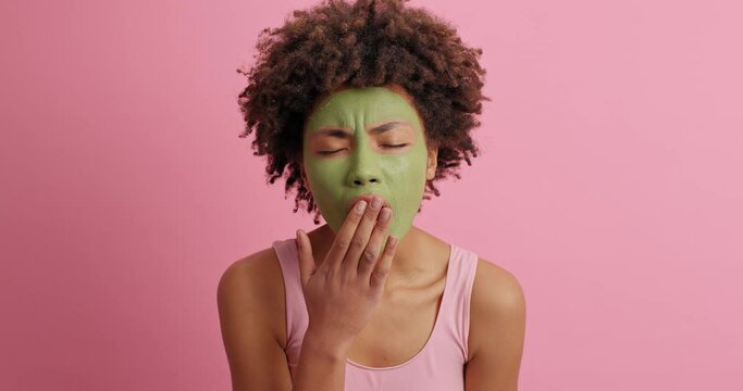 Morning beauty procedures concept. Serious tired young woman with Afro hair applies green facial mask yawns and covers mouth rejuvenates skin poses against pink background. Slow motion video