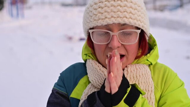 Portrait of a woman in glasses covered with hoarfrost. The girl is freezing and forgot gloves in very cold weather and blows on her bare hands