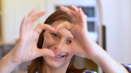 Smiling young woman showing hands sign heart shape looking at camera at home