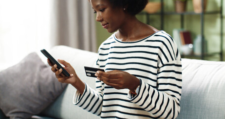 African American young happy beautiful woman sits on sofa in cozy living room in house tapping on...