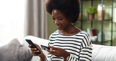 African American young happy beautiful woman sits on sofa in cozy living room in house tapping on...