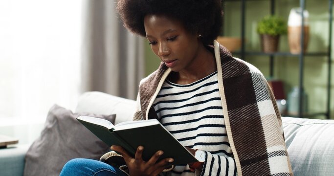 Pretty African American young happy female sitting on couch covered in plaid resting and reading favorite novel enjoying book at home. Leisure concept. Woman studying in cozy room. Self-education