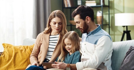 Portrait of joyful Caucasian young parents teaching small adorable child daughter typing, browsing online on tablet while sitting on sofa together at home. Family time concept. Tech gadget using