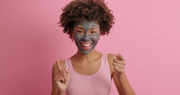 Positive curly African American woman wears black clay mask on her face moves carefree indicates at camera then raises arms and dances isolated over pink background. Self care and beauty concept
