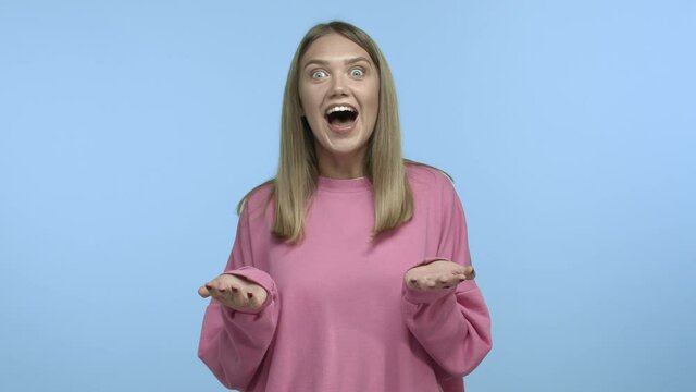 Slow motion of beautiful blond woman raising eyebrows surprised, spread hands sideways with disbelief and starts to laugh out loud from funny story, chuckle against blue background