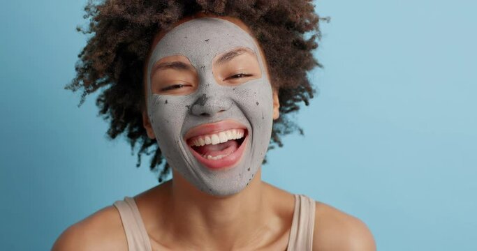 Positive female model with curly bushy hair smiles broadly has white perfect teeth bare shoulders wears clay mask on face cares about skin and complexion isolated over blue background takes day off