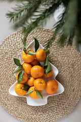 Small ripe tangerines with leaves, on plate in shape of Christmas tree on wicker serving napkin. Concept winter vitamin food. Moody still life festive composition. For greeting card, book, stories