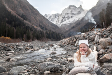 Little blond girl eight years old dressed in knitted warm hat, sweater and wrapping plaid blanket lies on the pillow against  the river landscape. Happy hiker wanderlust the view of snow, mountains