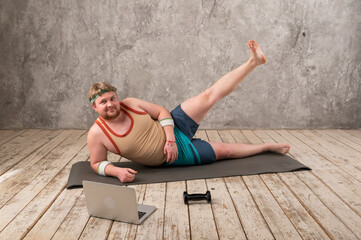 funny fat man in sports clothes doing exercises. He plays sports remotely while looking at his laptop