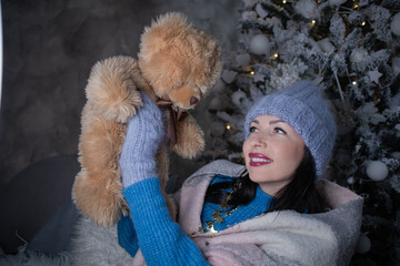 brunette in a blue sweater and blue hat and mittens with a teddy bear near the new year tree