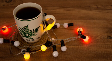 coffee mug with 'home sweet home' on it close-up and christmas decorations on wooden table background with colorful lights. Stay home new year. Top view and space for text