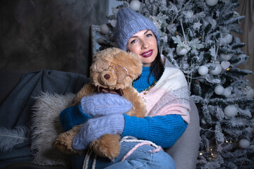brunette in a blue sweater and blue hat and mittens with a teddy bear near the new year tree