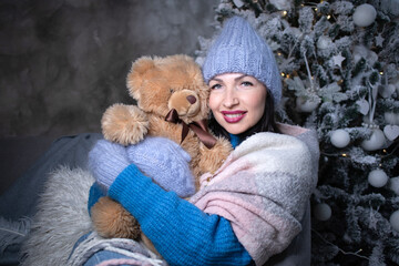 brunette in a blue sweater and blue hat and mittens with a teddy bear near the new year tree