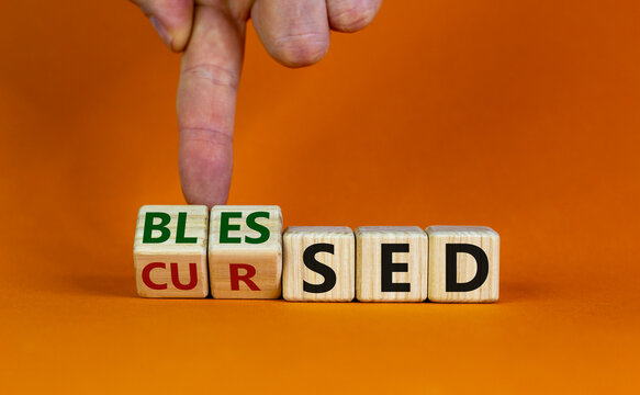 Cursed or blessed. Male hand flips a wooden cube and changes a word 'cursed' to 'blessed' or vice versa. Beautiful orange background, copy space. Religious and cursed or blessed concept.