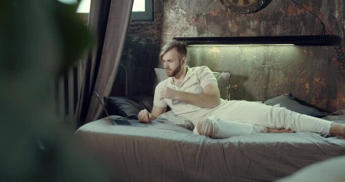 a man lying on the bed with his laptop, learning and working online. Slow motion