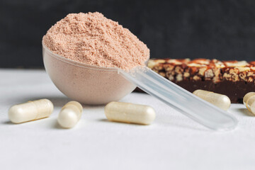 Scoop of whey or soy protein isolate, chocolate protein bar, white capsules of amino acids and creatine, bodybuilding food supplements on white table and dark background with copy-space