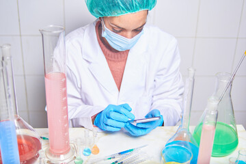 female chemist scientist with face mask using her mobile phone while working in the laboratory with...