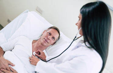 Fototapeta premium Beautiful young smiling confident woman doctor with a stethoscope is examining the senior patient in the hospital ward