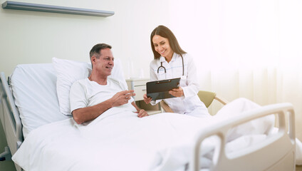 Handsome mature patient man is lying on the clinic bed and has a conversation with a confident young woman doctor in a white coat