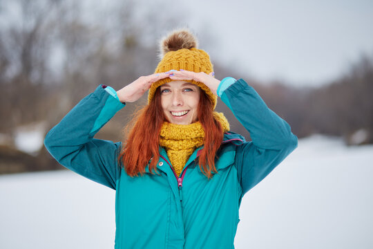 Happy smiling young woman with long red hair and clear skin looks into the camera with two hands on head. Girl is wearing an orange scarf and hat, and a blue ski suit