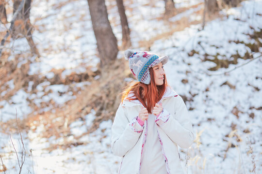 Girl with long red hair and a funny white-blue hat poses against the backdrop of a winter forest. Outdoor activities in the winter season