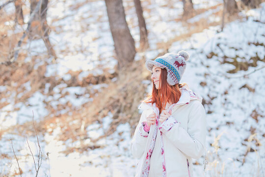 Girl with long red hair and a funny white-blue hat poses against the backdrop of a winter forest. Outdoor activities in the winter season
