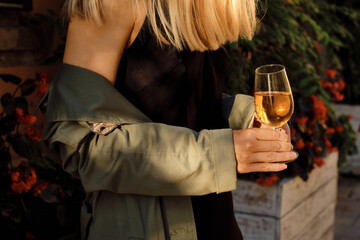 Beautiful girl in a trench coat and evening dress with a glass of wine. A girl walks in an empty morning city street at dawn after a night party. Enjoy the summer days. Celebrate the holiday alone