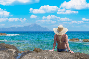 back view beautiful tan woman in swimsuit sitting on stone beach, with mount Athos on background