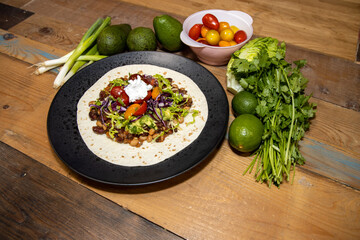 A plate of delicious smoky vegan chilli in a wrap and the ingredients used to make it on a wooden kitchen work top