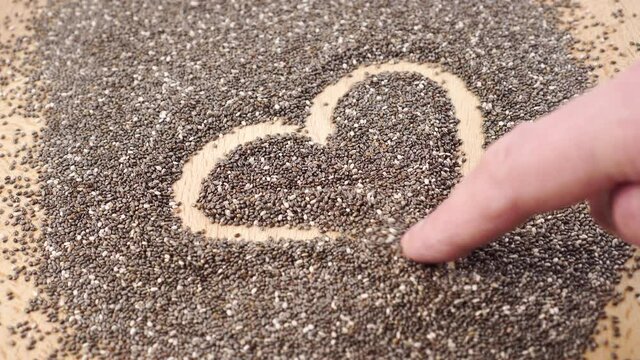 Hand is drawing a heart symbol on a heap of chia seeds. Close up. On a brown wooden surface. Romantic superfood and healthy food concept.