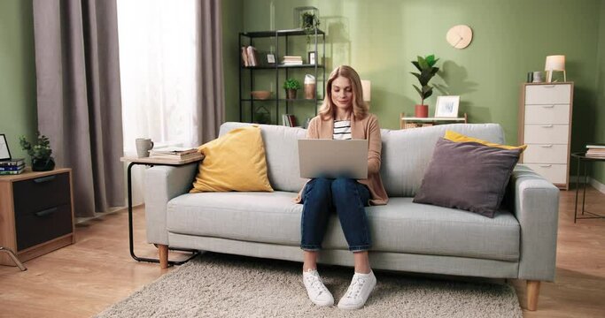 Caucasian beautiful young woman sitting on sofa in cozy living room in apartment texting and working on laptop. Distance work from home. Leisure concept. Browsing online on computer