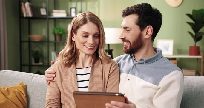 Close up portrait of happy smiling Caucasian married couple wife and husband searching internet, browsing online choosing and buying stuff together using tablet while sitting resting on sofa at home