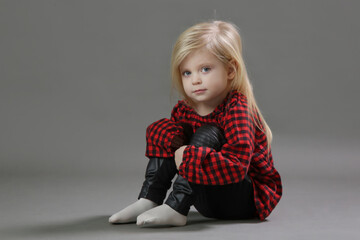 Fashion studio portrait of adorable 3 years old girl in red plaid shirt and leather black pants on grey background.