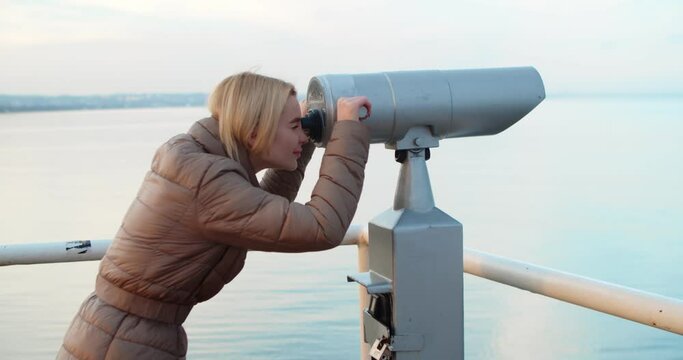
Pretty young woman on the road looks in a telescope on the sea pier in winter. Woman Enjoying the Seascape Looking Through a Telescope.