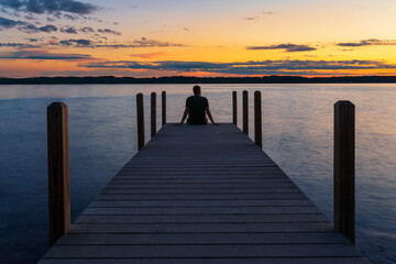 Man sitting on dock looking at sunset over lake.   - obrazy, fototapety, plakaty