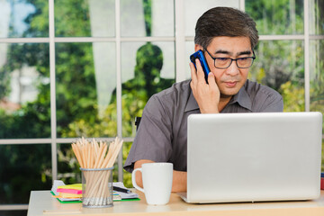 Asian executive senior businessman sitting on desk office he using his mobile phone and talking with somebody, the confident middle aged handsome man using laptop computer at workplace home office