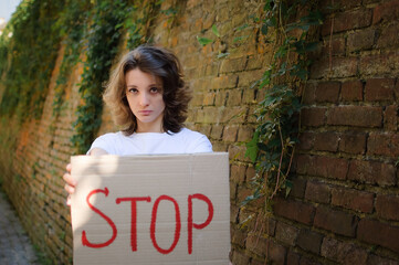 Young protesting woman in white shirt and jeans holds protest sign broadsheet placard with slogan 'Stop' for public demonstration on wall background.