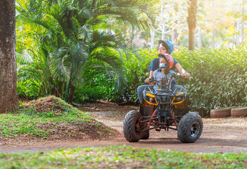 Asian mother and daughter riding quad bike in farm of Thailand, travel while covid-19, child has medical mask on face, medical mask with strap hang on neck. Background of green trees in farm. © dul_ny