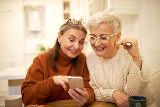 Two stylish retired female friends enjoying modern electronic gadget, sitting at cafe, watching pictures on mobile, recollecting good old days. Elderly women shopping online using smart phone