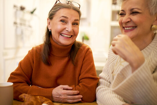 Indoor Image Of Two Joyful Positive European Female Friends On Retirement Having Fun, Talking, Telling Funny Jokes During Meeting At Home Or Cafe. People, Friendship, Age And Retirement Concept