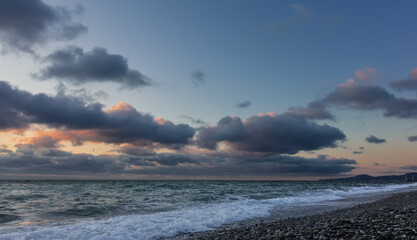 At sunset, blue clouds stand out in pink. The surf leaves an openwork white foam on the beach pebbles. The outline of the cape is visible on the horizon. Black Sea