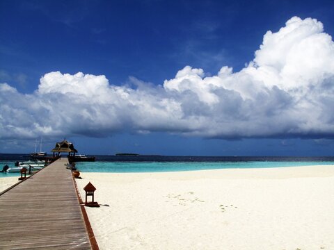 Beautiful sand beach with a wooden path leading to the pier, out to the blue ocean, on a sunny but partly cloudy day.
