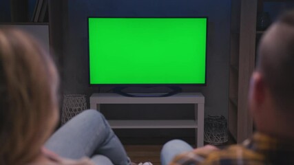 Family Couple Watches Green Screen TV Mock-up Sitting on Couch in Living Room Together. Rear View on Casual People who Watching TV Green Screen in Domestic Cinema. Looking TV Show or News in Home Rest - Powered by Adobe
